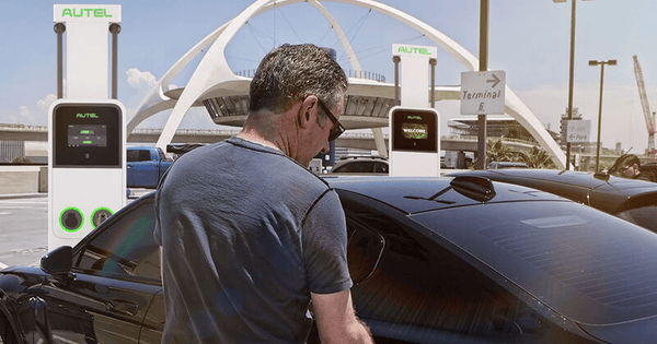 Man charging electric car at a charging station in front of a distinctive architectural structure under a clear sky.
